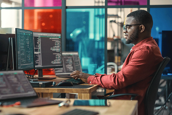 A Black male student sits at a desk in a computer lab. Two computer monitors are on a brown desk in front of him. The student has short black hair and matching facial hair. He wears black glasses and a red long-sleeved dress shirt. He sits in a black desk chair, typing on a black keyboard. Brown desks with computer monitors are also visible in the lab. Blue, red, purple, pink, and clear-colored glass windows are in the background. 